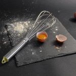Ovos, farinha e um batedor dispostos sobre uma placa de ardósia durante uma preparação culinária.