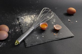 Ovos, farinha e um batedor dispostos sobre uma placa de ardósia durante uma preparação culinária.