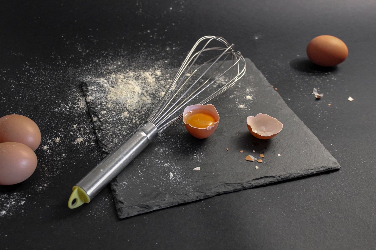 Ovos, farinha e um batedor dispostos sobre uma placa de ardósia durante uma preparação culinária.