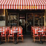 Café parisiense com toldo listrado vermelho e mesas externas vazias.