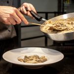 Chef finaliza um prato de massa cremosa com cogumelos em uma cozinha.