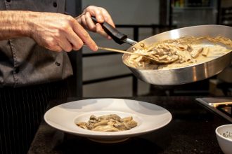 Chef finaliza um prato de massa cremosa com cogumelos em uma cozinha.